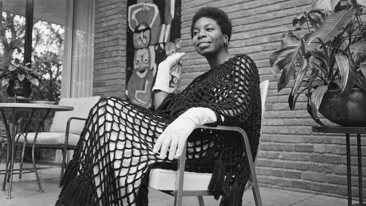 Nina Simone sitting in a chair wearing a black crocheted poncho and white gloves.