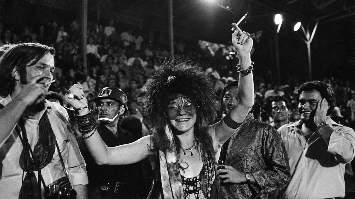 Janis Joplin smiling with her arms raised, holding a cigarette, surrounded by a crowd at an outdoor concert.