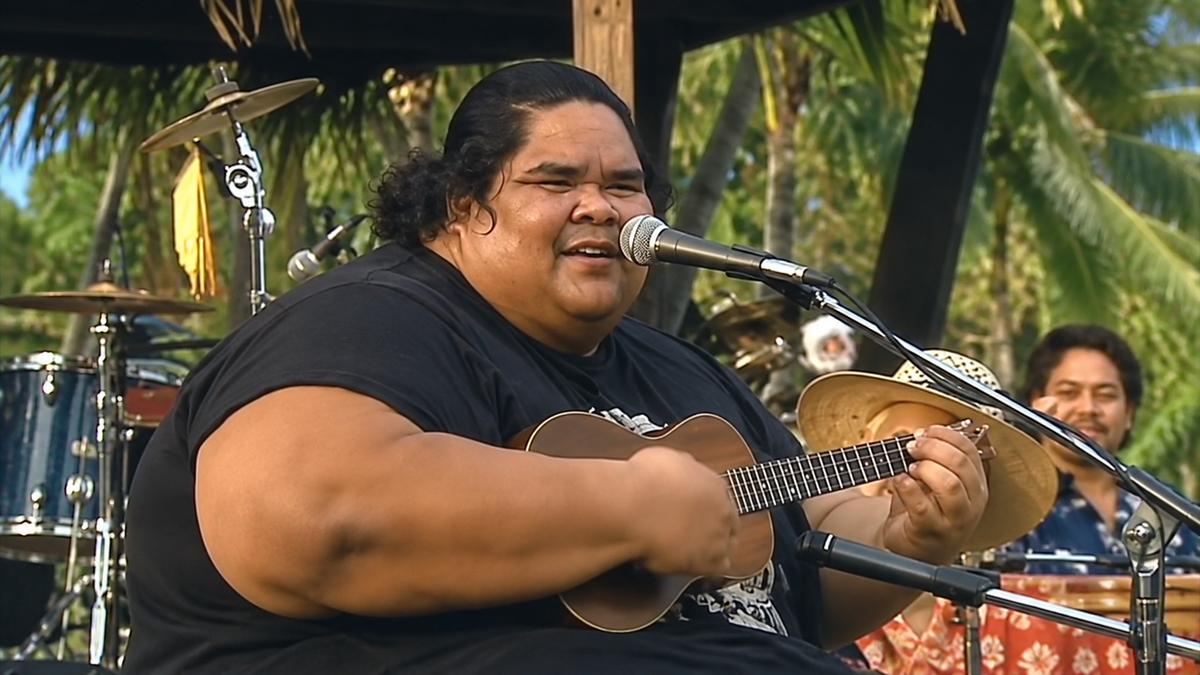 Israel Kamakawiwoʻole playing the ukulele and singing into a microphone on an outdoor stage.