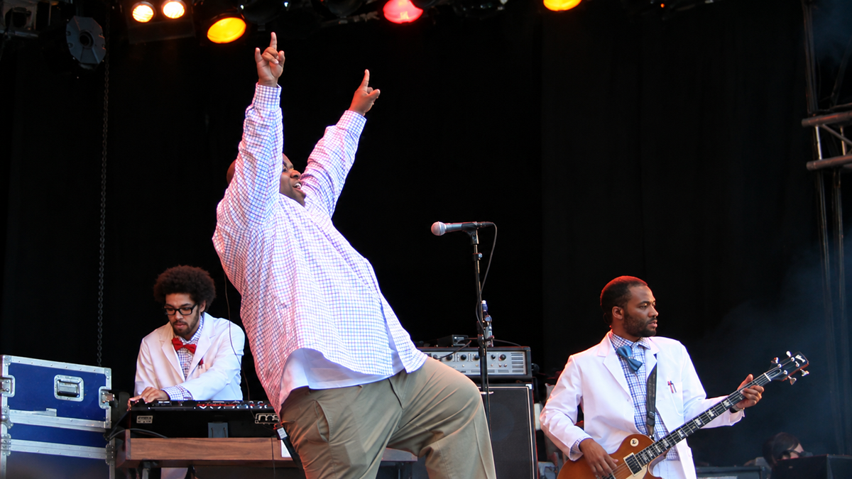 A singer in a plaid shirt with arms raised performs on stage with a keyboardist and a bassist, all wearing white jackets.