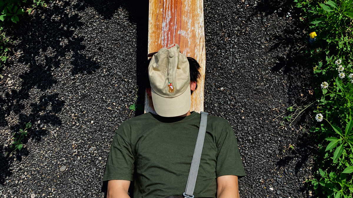 A person in an olive green t-shirt and a beige baseball cap lying on a wooden bench outdoors.