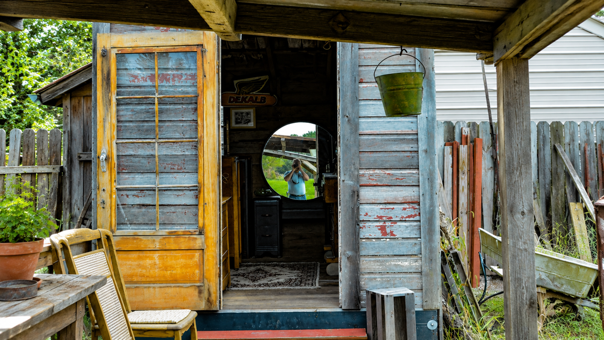 An open door leads into a weathered shed containing a round mirror that reflects a person taking a photo.