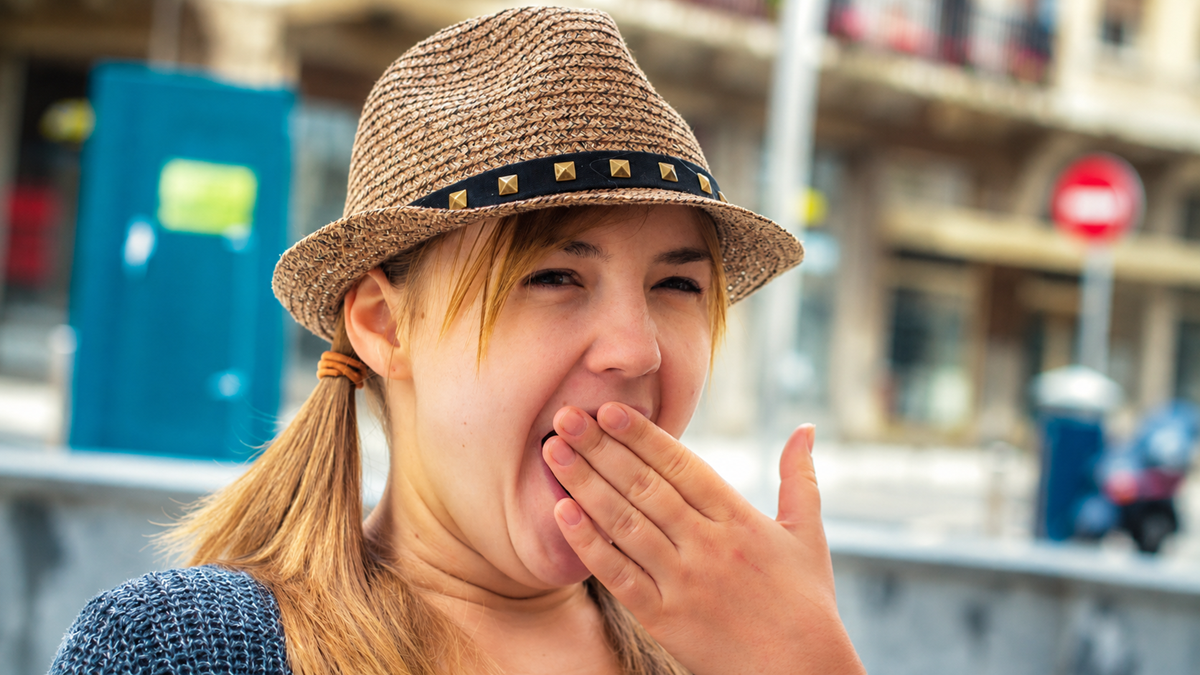 A young woman wearing a straw fedora with a studded band yawns while covering her mouth with her hand.