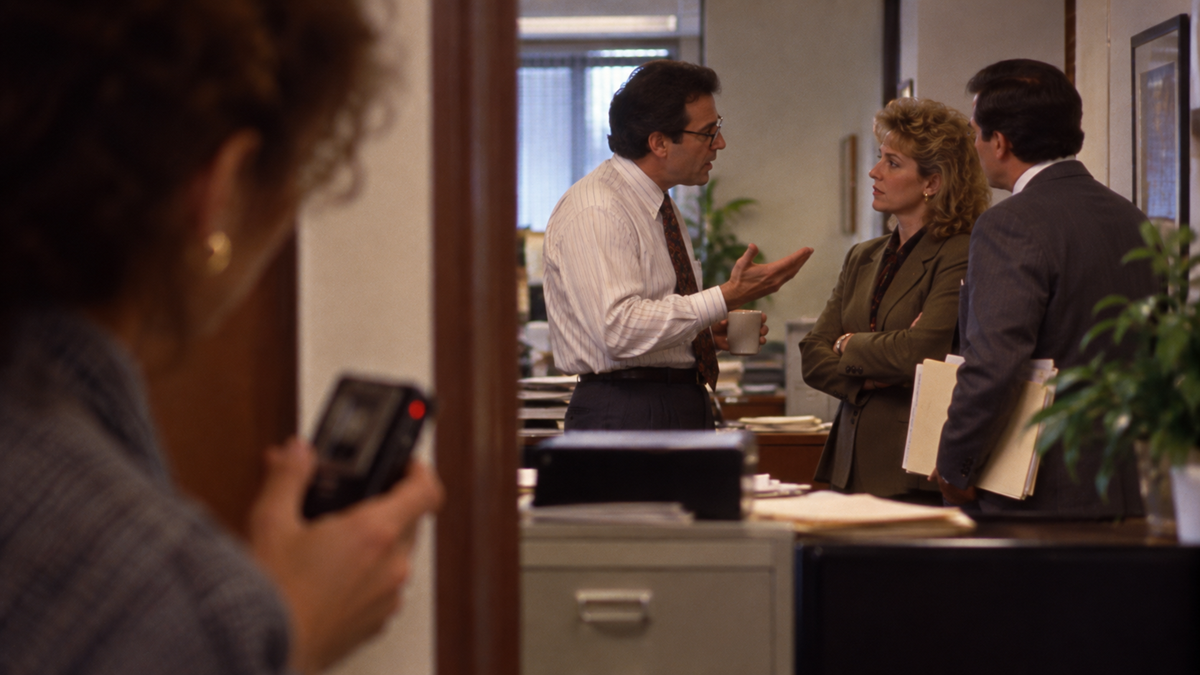 A woman in the foreground holds a camera, looking toward two men and a woman in business attire talking in an office.