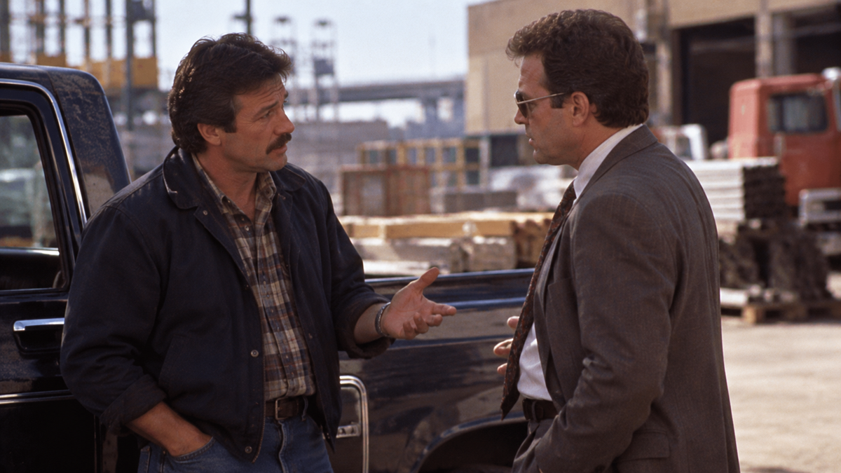 Two men in a parking lot, one wearing a jacket and plaid shirt and the other in a suit and sunglasses, face each other in conversation.