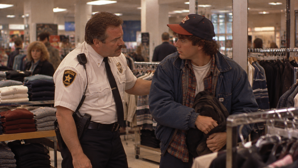 A security guard in a uniform puts a hand on the shoulder of a young man wearing a baseball cap and denim jacket inside a clothing store.