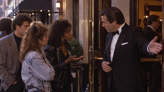 A man in a tuxedo gestures toward an entrance as he speaks with two women standing on the sidewalk.