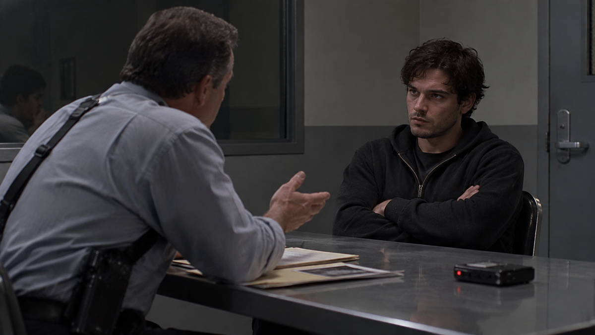 A man in a button-down shirt sits across from a younger man in a hoodie at a metal table in an interrogation room.
