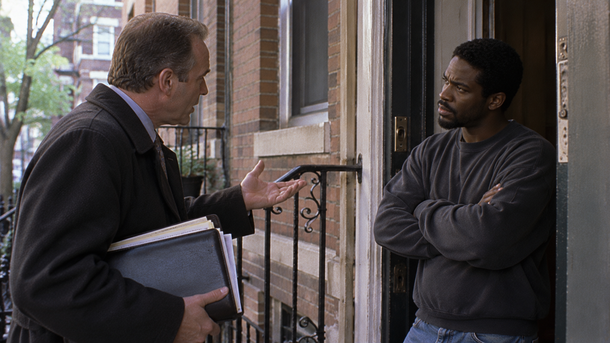 A man in a coat holding a folder gestures while talking to a man standing in a doorway with his arms crossed.