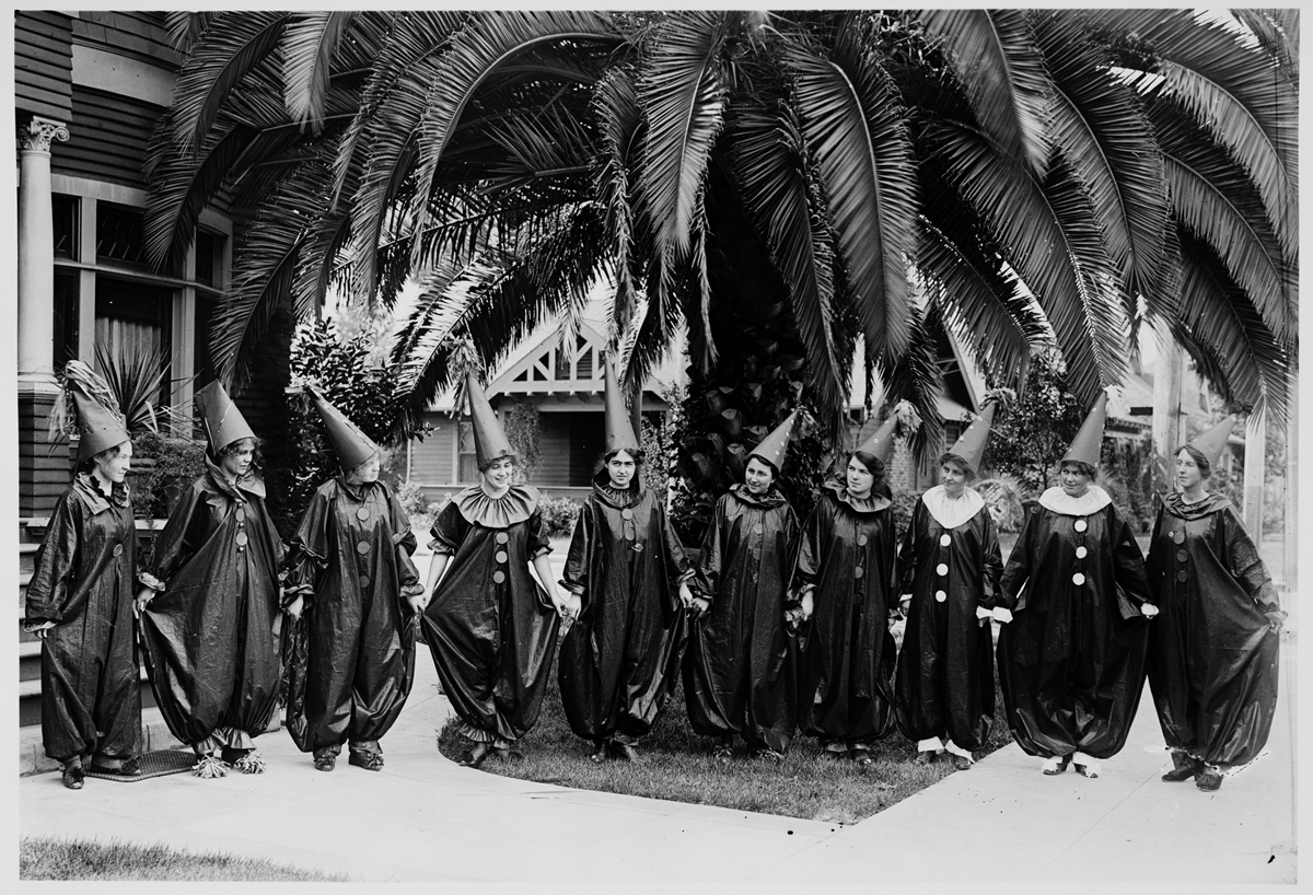 Nine people wearing identical dark, cone-shaped hats and loose, full-length costumes with large collars stand in a row in front of a palm tree.