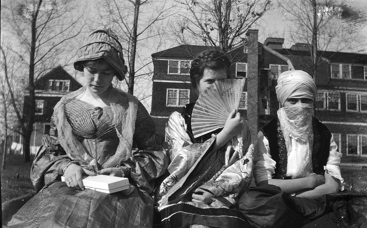 Three women in vintage costumes sit outdoors, with one holding a folding fan to cover her face and another wearing a head covering and face veil.