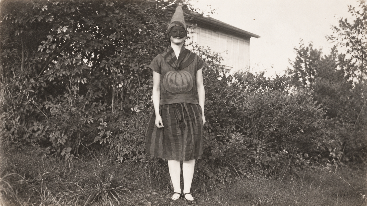 A person in a Halloween costume consisting of a pointed cap, a dark mask, and a striped dress featuring a pumpkin graphic on the chest.
