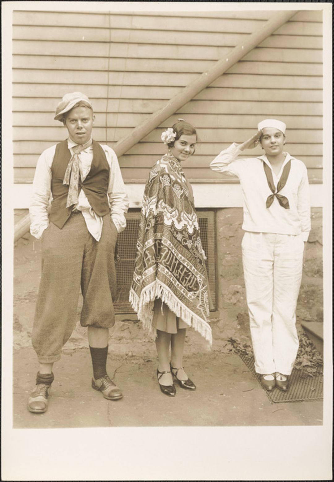 A black-and-white photo of three people: a man in a vest and cap, a woman wearing a patterned fringed shawl, and a person in a sailor uniform saluting.