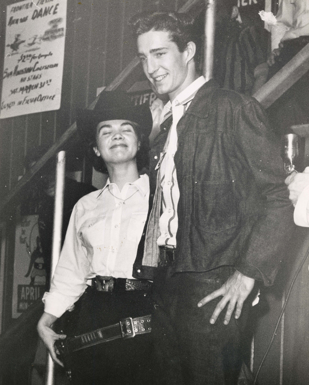 A young woman in a cowboy hat and a young man in a collared shirt and jacket standing together in front of a dance event poster.