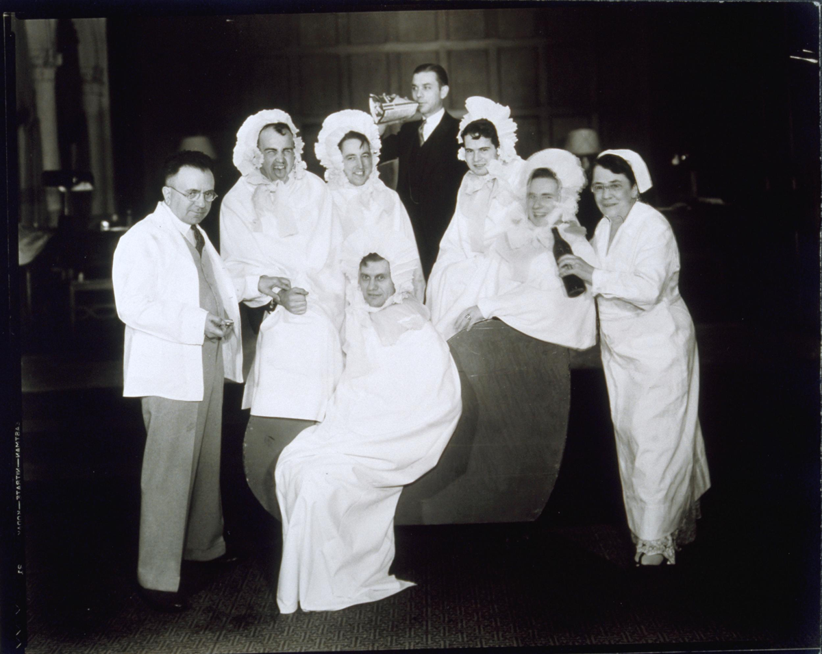 A group of men and women in nightgowns and nightcaps posing for a photograph, with one man standing in a suit and a man in the center playing a trumpet.