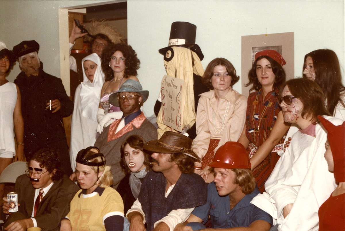 A group of people wearing diverse Halloween costumes, including a person dressed as a ghost with a sign, a person in a top hat, and others in casual or character outfits.