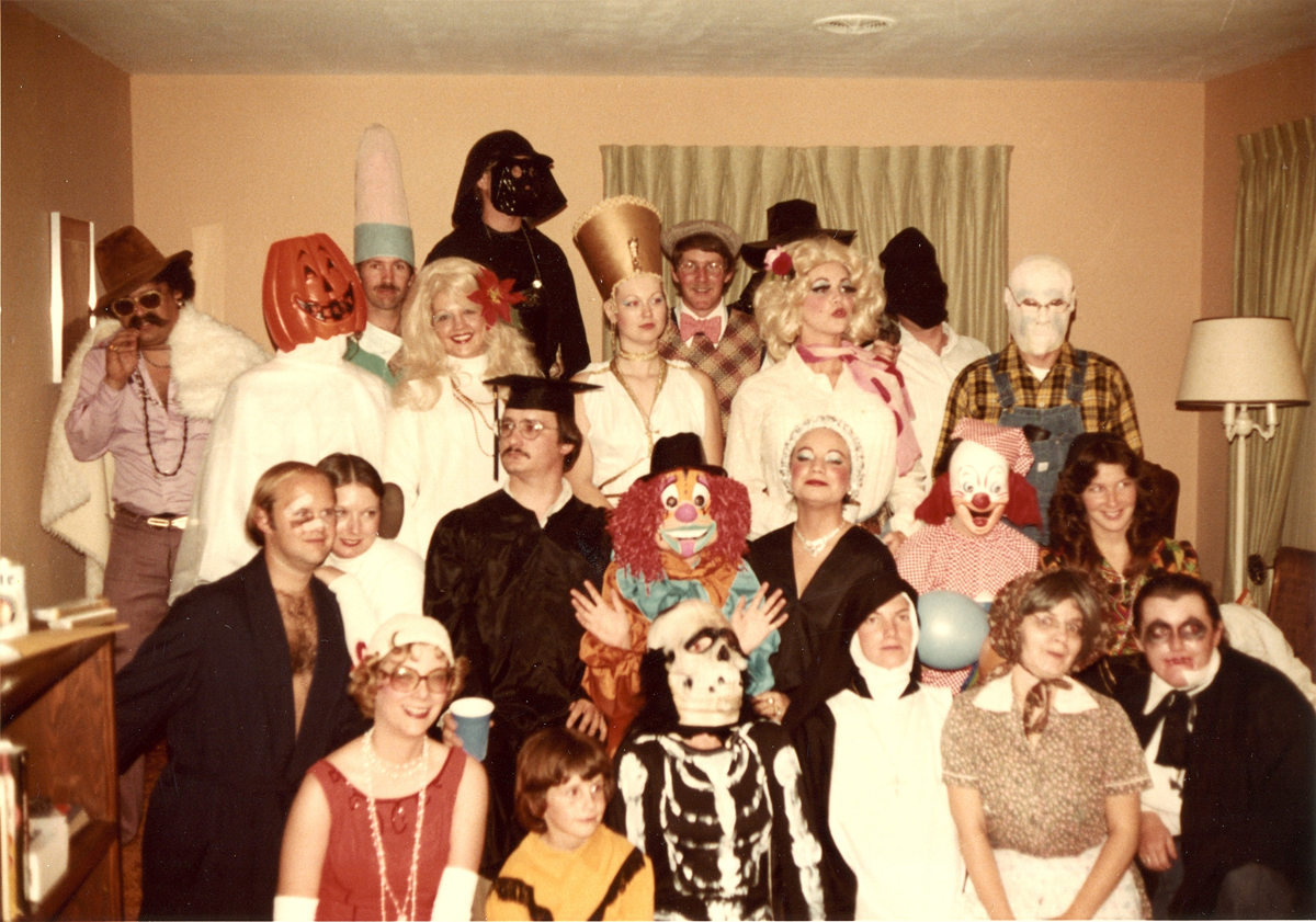 A large group of people in diverse Halloween costumes pose for a photo in a living room.
