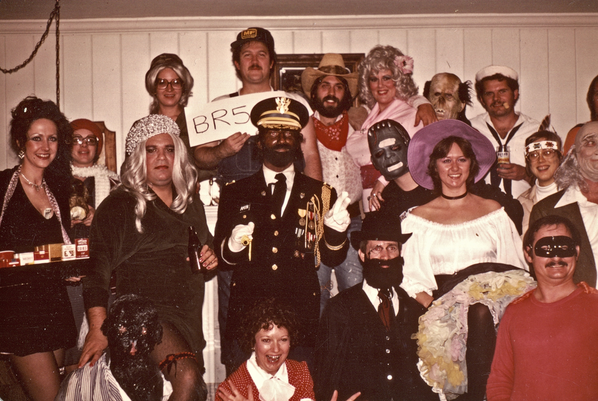A group of people wearing diverse Halloween costumes, including a person in a naval officer uniform holding a sign that says “BR549,” posing for a group photo indoors.