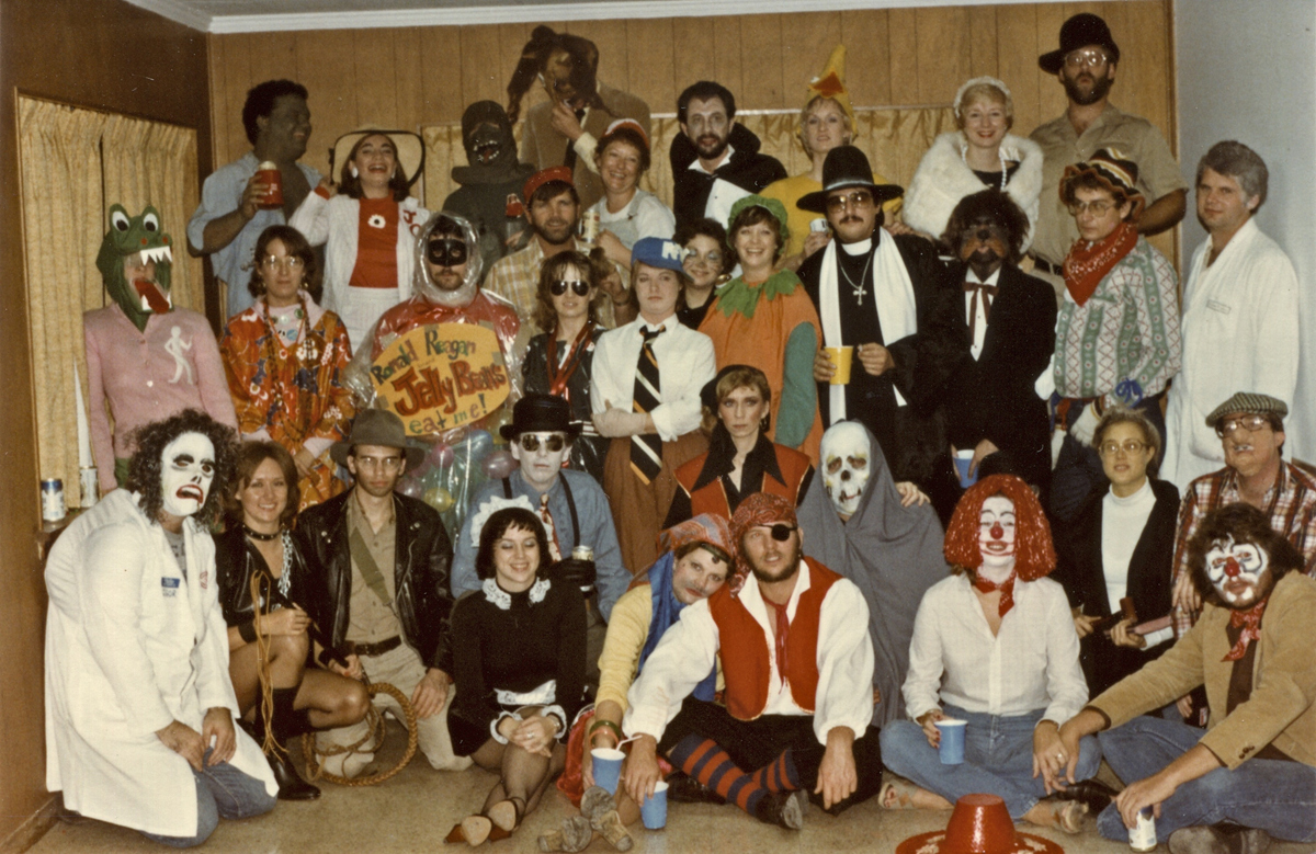 A large group of people in diverse costumes, including a Ronald Reagan jelly bean bag, clowns, and various historical figures, posing for a group portrait at a party.