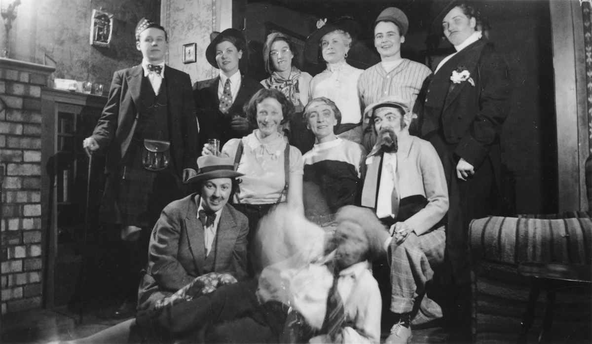A group of eleven people posing for a black-and-white group portrait, many wearing hats, ties, or theatrical costumes.