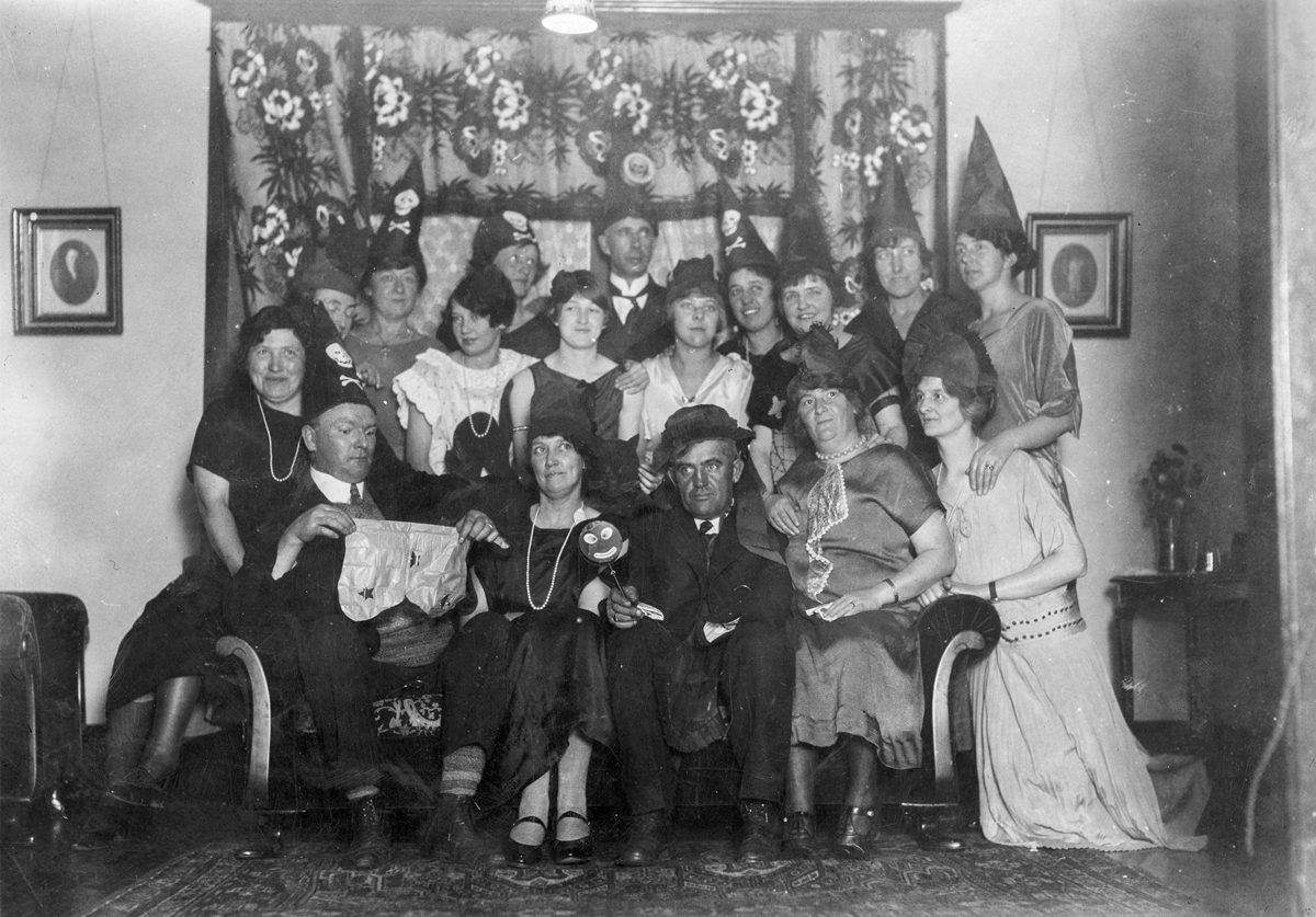 A group of men and women posed for a portrait, many wearing pointed party hats, with one man holding a pair of baby bloomers and another person holding a small mask on a stick.