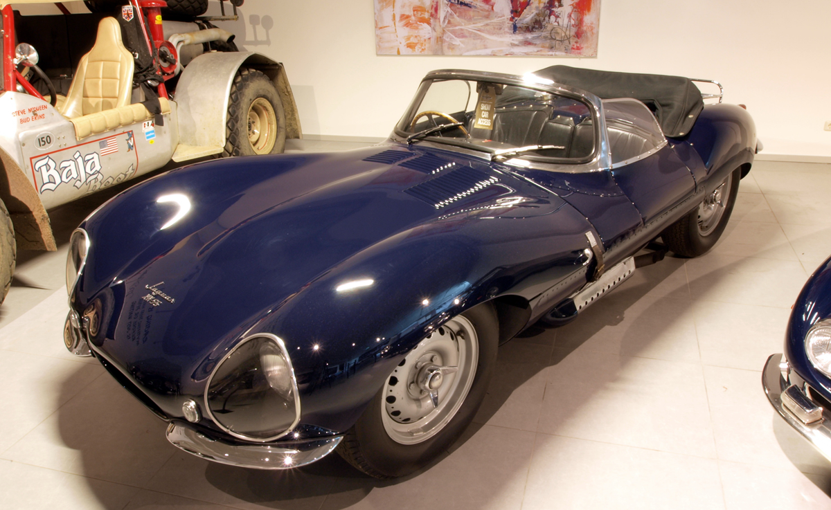 A vintage dark blue Jaguar XKSS sports car on display in a museum next to a Baja Boot off-road vehicle.