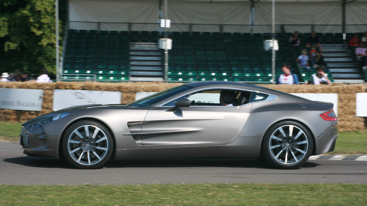 A grey Aston Martin One-77 drives along a track in front of bleacher seating.