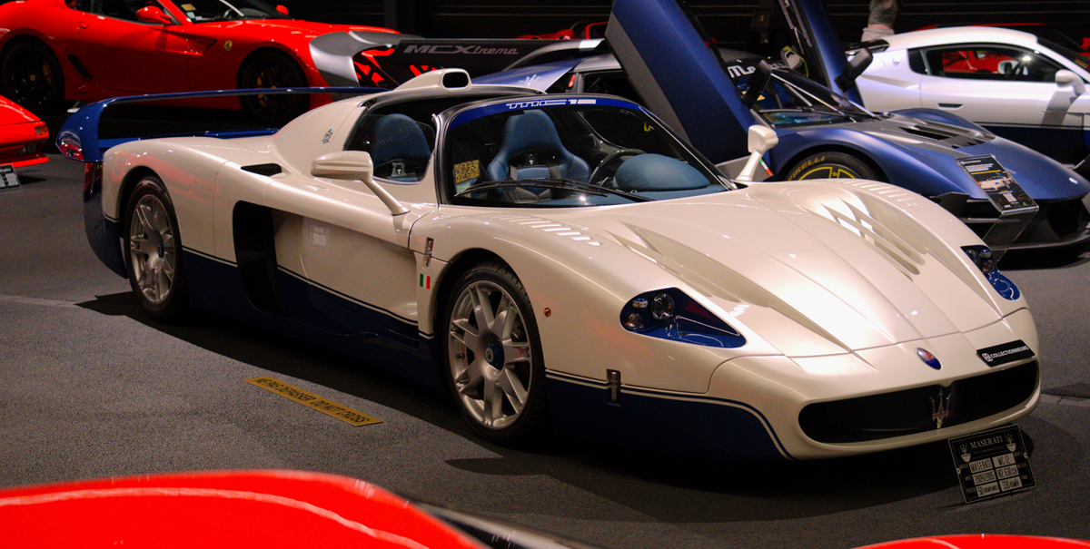 A white and blue Maserati MC12 sports car displayed on a museum floor with other luxury vehicles in the background.