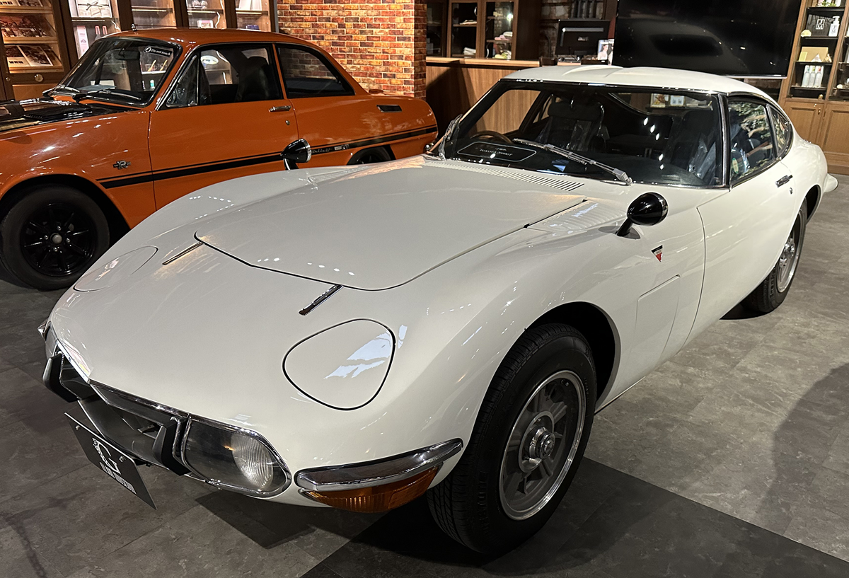 A white vintage Toyota 2000GT sports car parked next to an orange vintage car in a showroom.