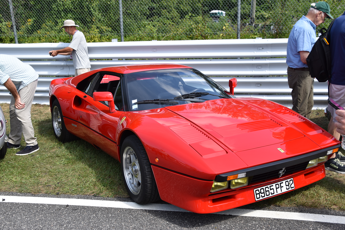 A red Ferrari 288 GTO parked on grass next to a trackside fence, with three people standing nearby.