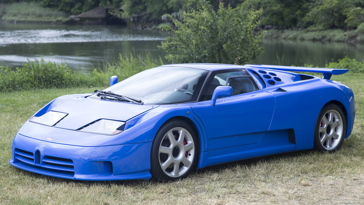 A bright blue Bugatti EB110 sports car parked on grass near a lake.
