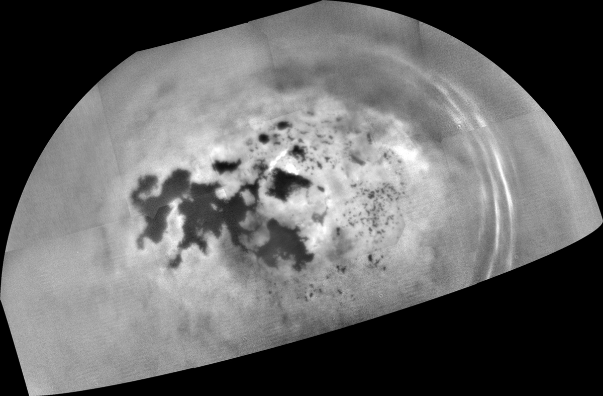 Black and white, semicircular mosaic image of a planetary surface showing a large, dark, irregular feature surrounded by bright, textured terrain and faint, concentric rings near the edge.