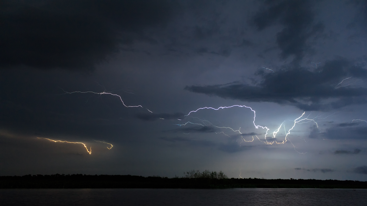 Lightning bolts illuminate a dark, cloudy sky over a body of water.