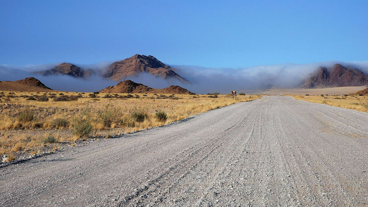 A gravel road leads through a dry landscape toward mountains partially obscured by low-hanging clouds.
