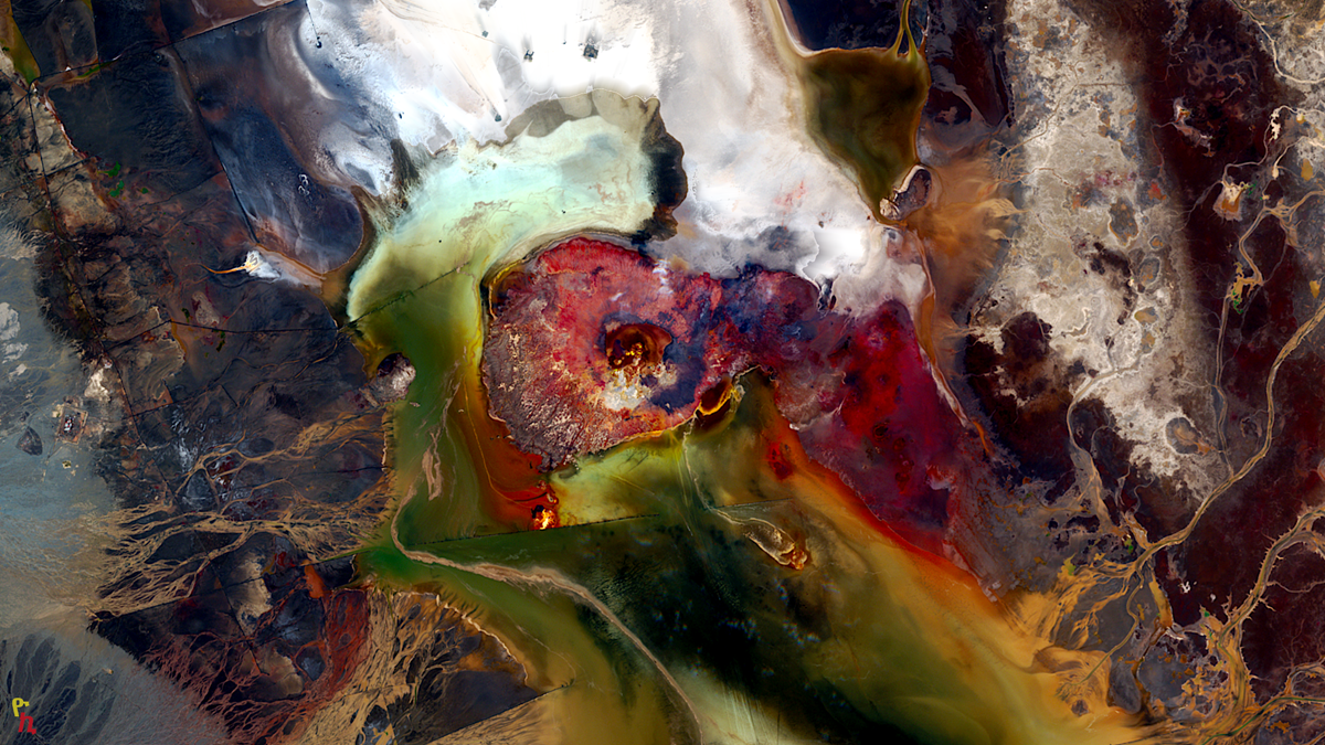 Satellite view of a mineral-rich lake with vibrant, swirling patterns of red, white, green, and brown pigments.