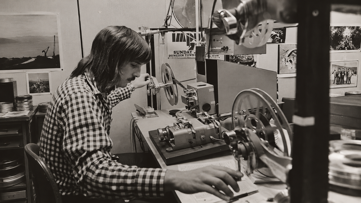A man with long hair in a checkered shirt operates a film editing machine in an office.