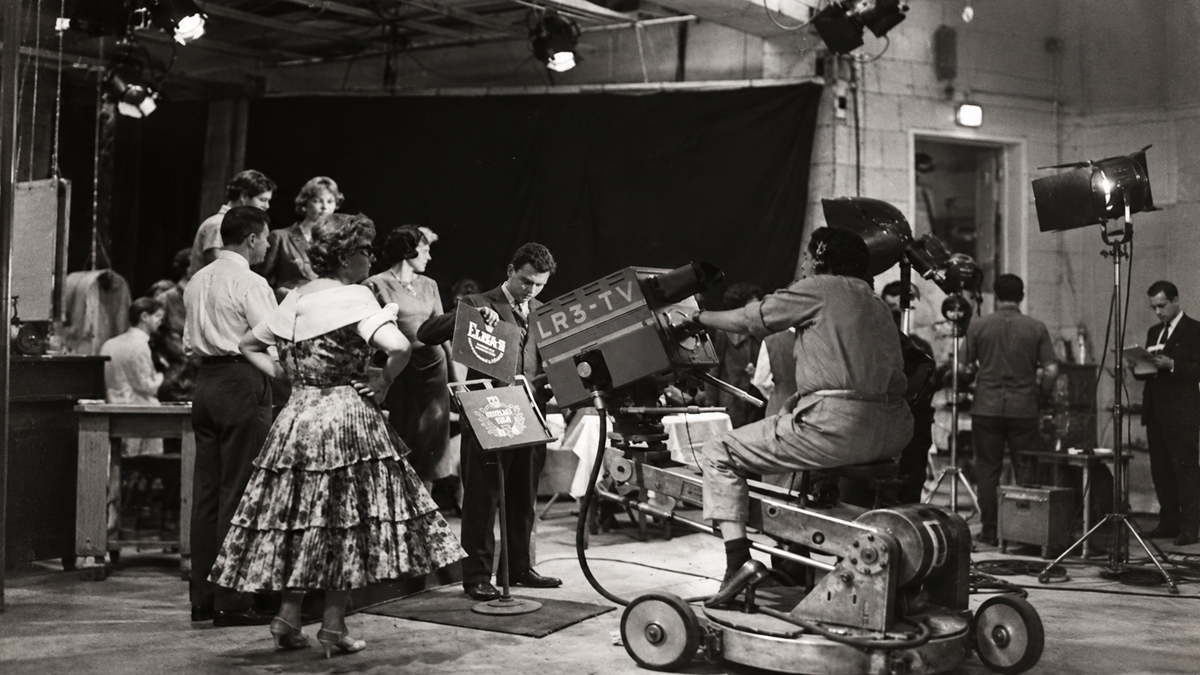 A television studio scene with a cameraman filming a group of people standing on a set.