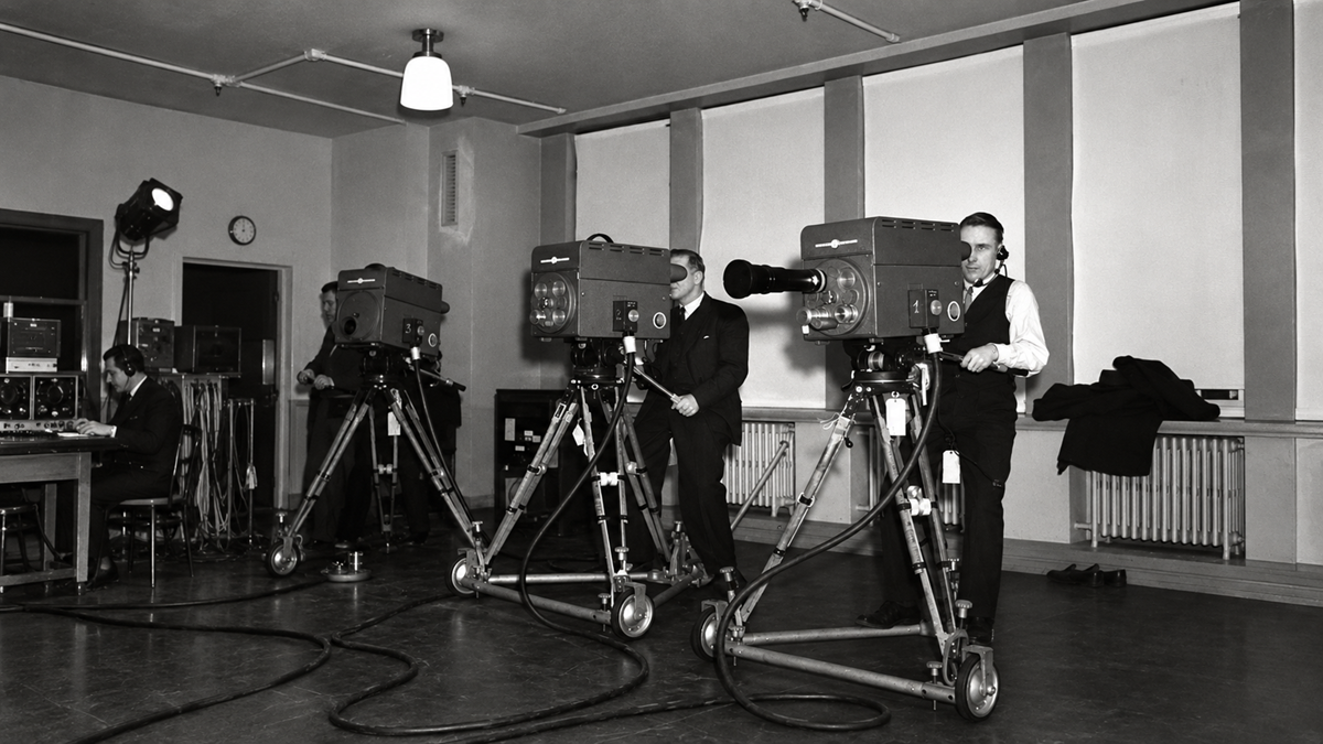 Three camera operators work in a television studio, each standing behind a large vintage camera on a rolling tripod.