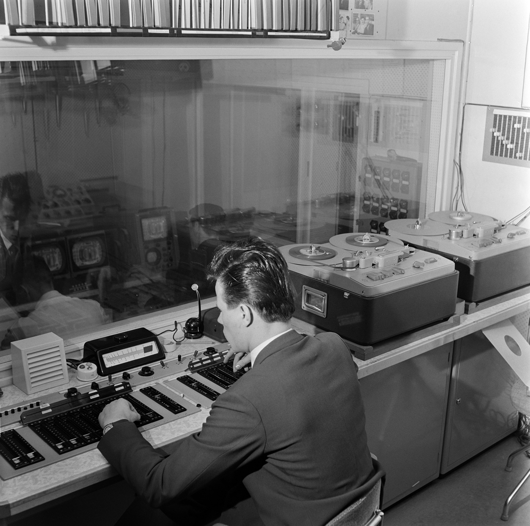 A man in a suit operates an audio mixing console in a radio control room, with reel-to-reel tape recorders on the desk beside him.