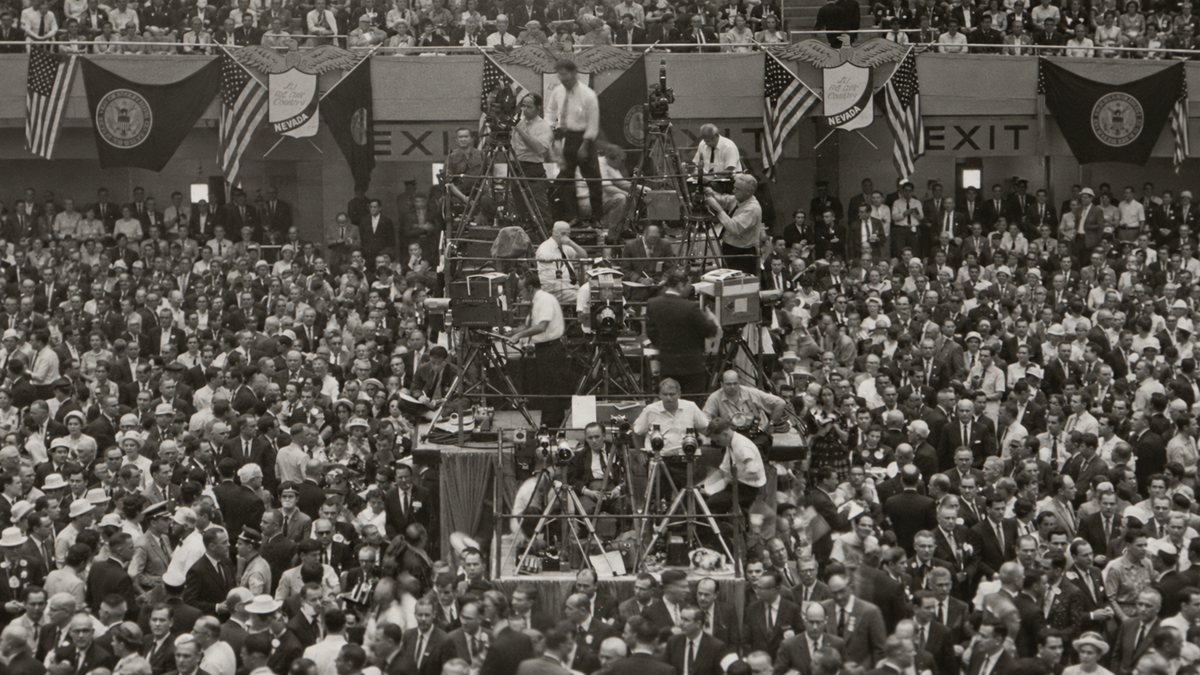 A crowded political convention hall with multiple television camera crews stationed on a central raised platform.