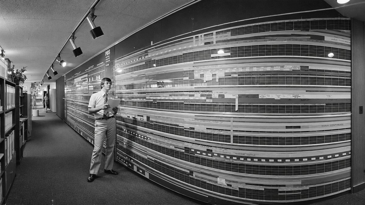 A man holding a document stands in an office hallway next to a long, curved wall covered with a large, detailed broadcast programming schedule.