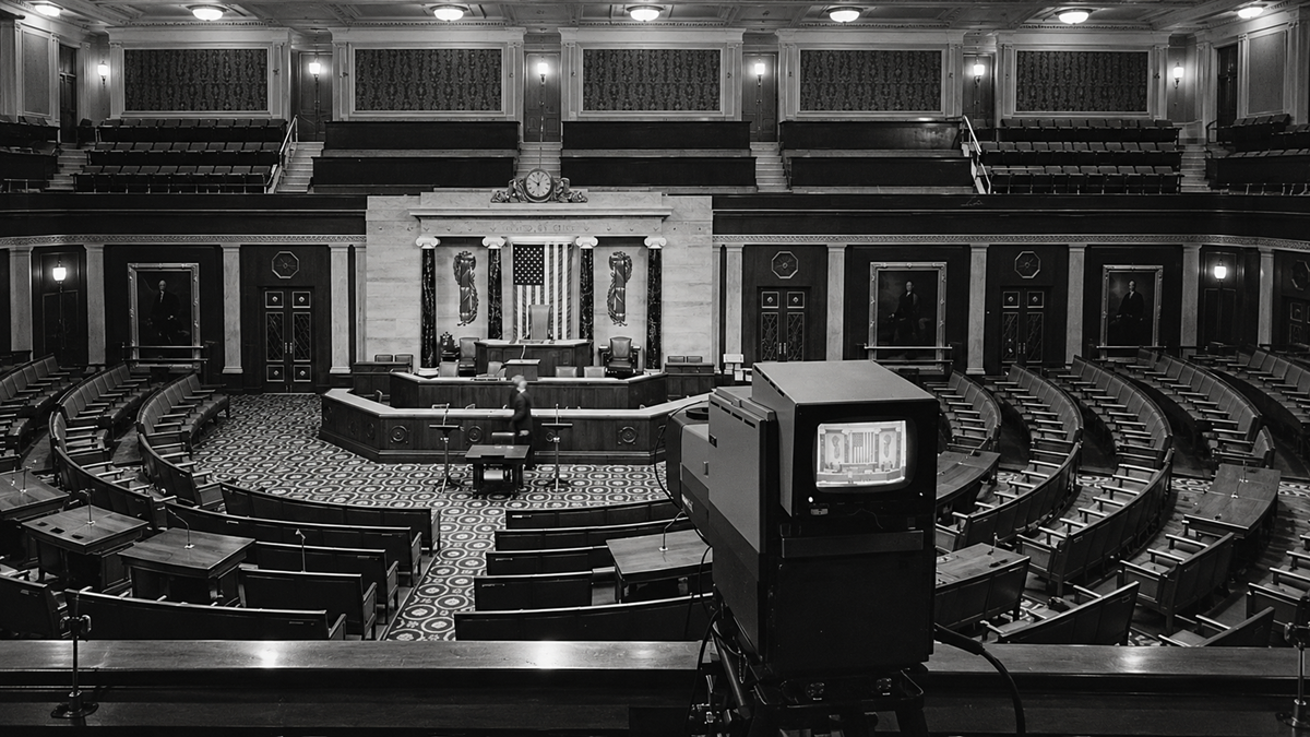 A black-and-white view of the U.S. House of Representatives chamber from behind a television camera, showing rows of empty desks and a central podium.