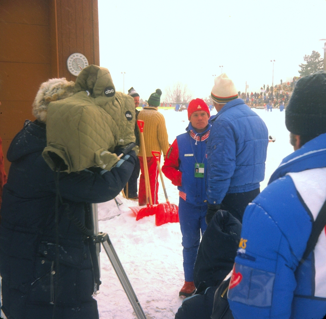 A camera operator films a person in a red and blue athletic uniform standing on snow, surrounded by spectators and people in winter gear.