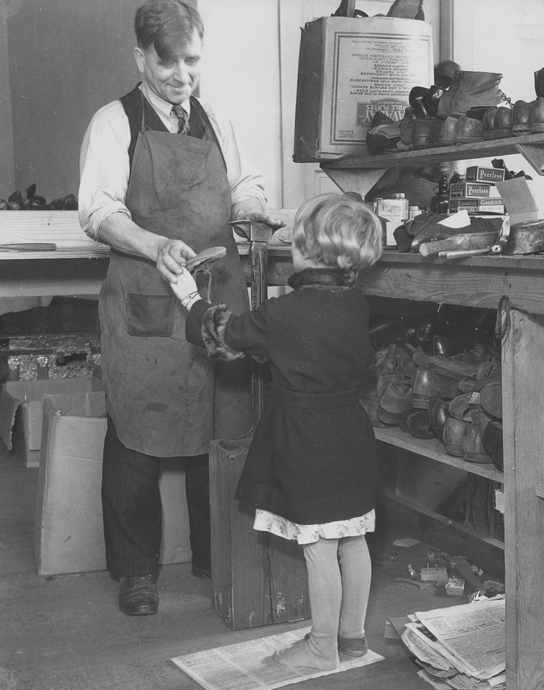 A cobbler in an apron measures the foot of a young girl standing in his shop.