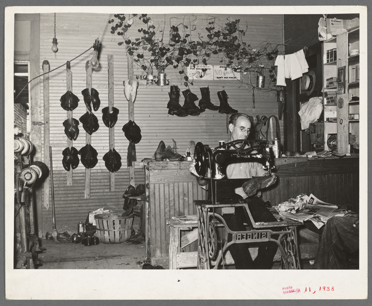 A cobbler working at a Singer sewing machine in a shop filled with hanging shoes and plants.