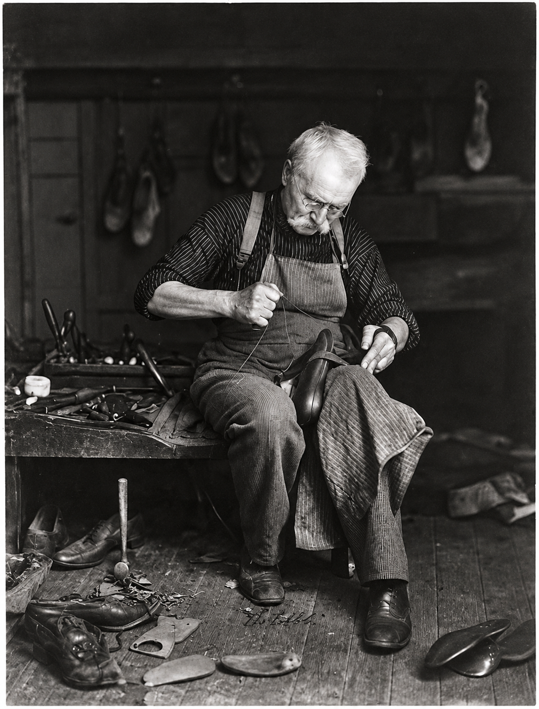 An elderly cobbler with glasses and a mustache uses a needle and thread to repair a leather shoe on his lap in a workshop.