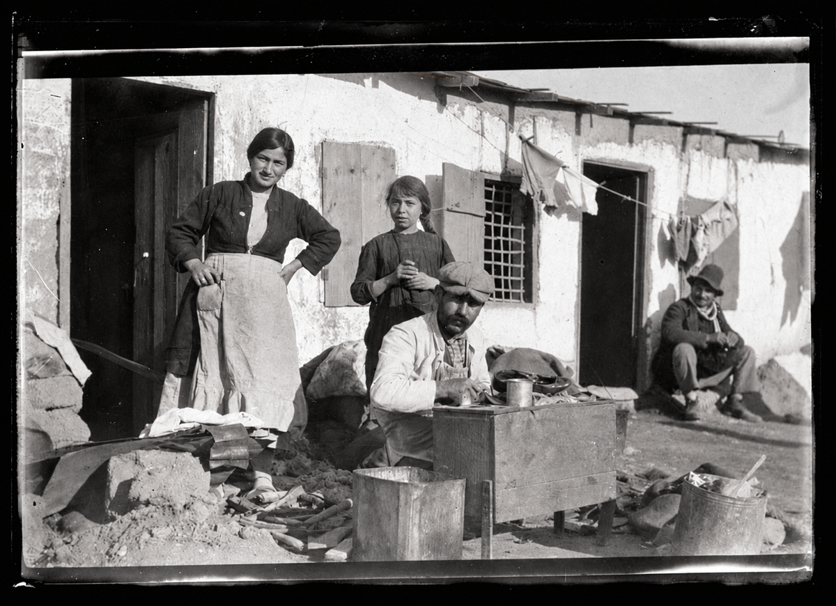 A man works at a small wooden bench while a woman and a girl stand behind him in front of a white building, with a man sitting on the ground nearby.