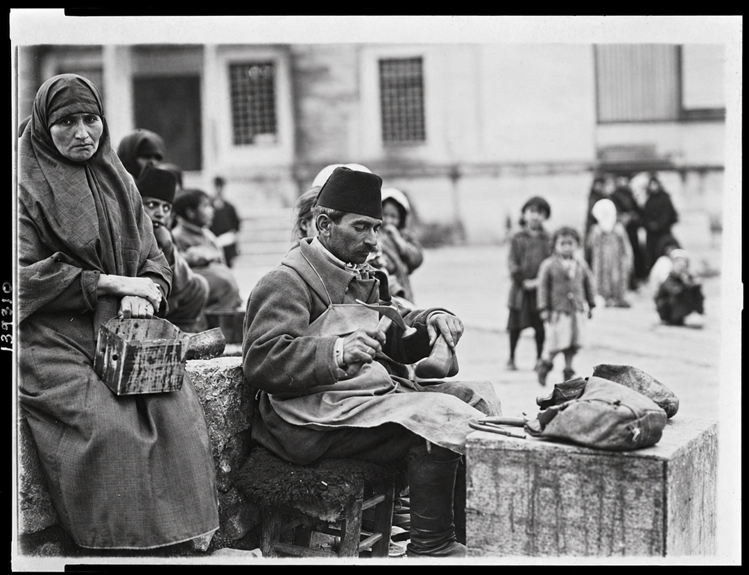 A man in a fez sits on a stool cobbling a shoe, while a woman in a headscarf sits beside him holding a wooden box in a public square.