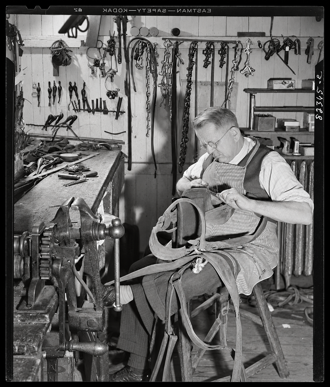 An elderly man wearing glasses and an apron sits on a stool in a workshop, stitching a leather harness.