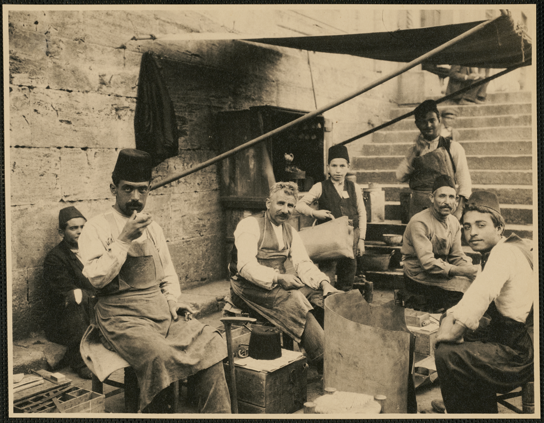 Six men and boys wearing fezzes work together in an outdoor workshop with stone walls and stairs.